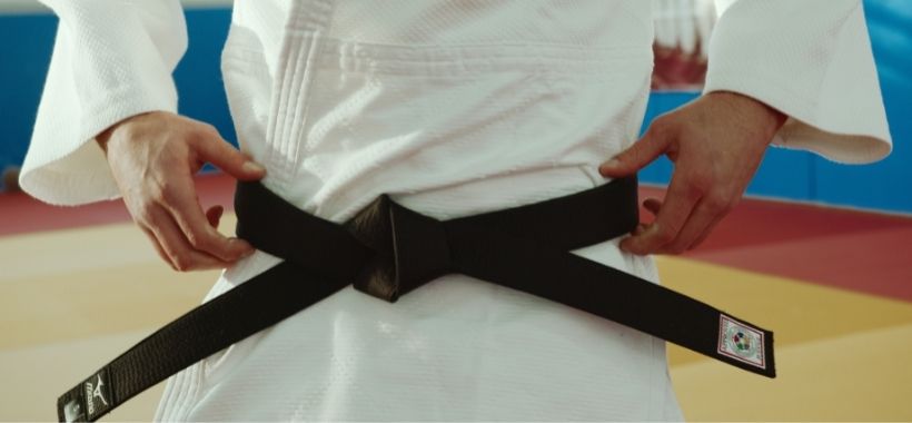 Fotografia fragmentada mostrando uma pessoa usando um kimono com a faixa na cor preta. As mãos estão segurando a faixa, que está presa à cintura.