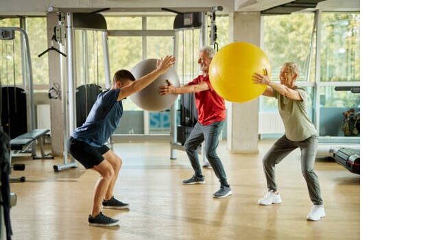 Senior Couple Exercising With Personal Trainer Using Fitness Ball At The Gym