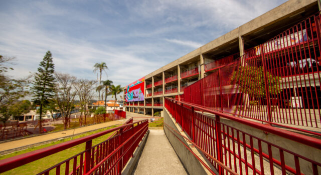 A imagem mostra o CEU São Pedro/José Bonifácio, um prédio de concreto com grades vermelhas e um grande mural colorido na fachada. Uma rampa com corrimãos vermelhos leva até o edifício. Ao lado, há árvores e um gramado sob um céu azul com nuvens finas.