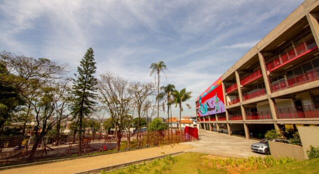 A foto mostra a fachada do CEU São Pedro/José Bonifácio em um dia ensolarado. À direita, aparece um prédio de três andares com grades vermelhas e um grande mural colorido na lateral. À esquerda, há árvores de diferentes tipos e alturas, além de parte do bairro ao fundo. Em primeiro plano, vê-se um gramado com pequenas plantas e uma área pavimentada próxima ao prédio. O céu está azul com nuvens finas.