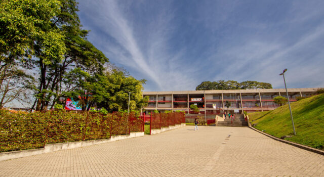 Uma foto externa do CEU São Pedro/José Bonifácio, mostrando uma ampla área pavimentada levando até um prédio escolar de dois andares. Há jardins, árvores e cercas vermelhas ao redor, sob um céu azul com poucas nuvens.