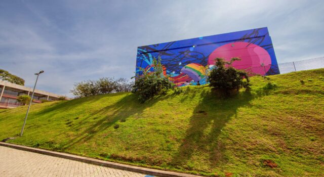 A foto mostra uma área externa ensolarada, com um gramado verde em uma ladeira inclinada. No topo da ladeira há um grande painel colorido na fachada de um prédio — o CEU São Pedro/José Bonifácio. O painel exibe um mural vibrante, com formas abstratas em cores fortes como azul, rosa, roxo, amarelo e verde, criando um visual dinâmico. Em frente ao mural há alguns arbustos e pequenas árvores que projetam sombras longas sobre o gramado. Na parte inferior da imagem, vê-se um caminho pavimentado com blocos de concreto. À esquerda, parte da estrutura do prédio aparece ao fundo, junto a um poste de iluminação. O céu está claro, com poucas nuvens, indicando um dia ensolarado.