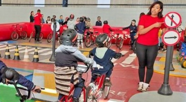 Crianças andando de bike, em uma pista com sinais de trânsito, acompanhadas por monitoras.